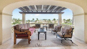 A covered outdoor seating area with a sofa, chairs, and a table, overlooking a coastal landscape.