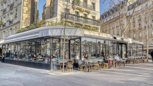 Un café al aire libre con paredes de vidrio, muebles de mimbre y un área de estar cubierta.