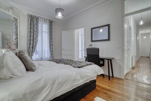 A bedroom with a large bed, a desk, and a chair. There is a window with curtains, a door, and a hallway leading to another room. - [120 m2] Luxury Eiffel Signature (Paris)
