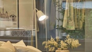 Un dormitorio con una cama, una lámpara de noche, una ventana con vista a un edificio y ropa colgada afuera.