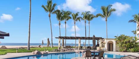 A pool area with lounge chairs and umbrellas, surrounded by palm trees and a view of the ocean.