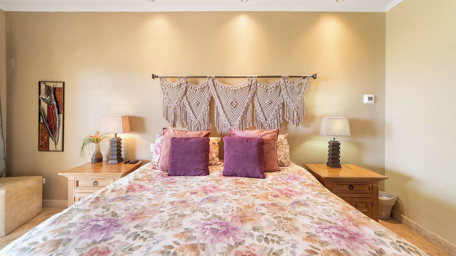 A bedroom with a floral bedspread, two purple pillows, a macrame wall hanging, bedside lamps, a small plant, and a framed artwork on the wall.