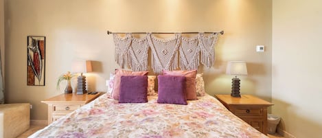 Un dormitorio con cubrecama floral, dos almohadas moradas, un tapiz de macramé, lámparas de noche, una planta pequeña y una obra enmarcada en la pared.