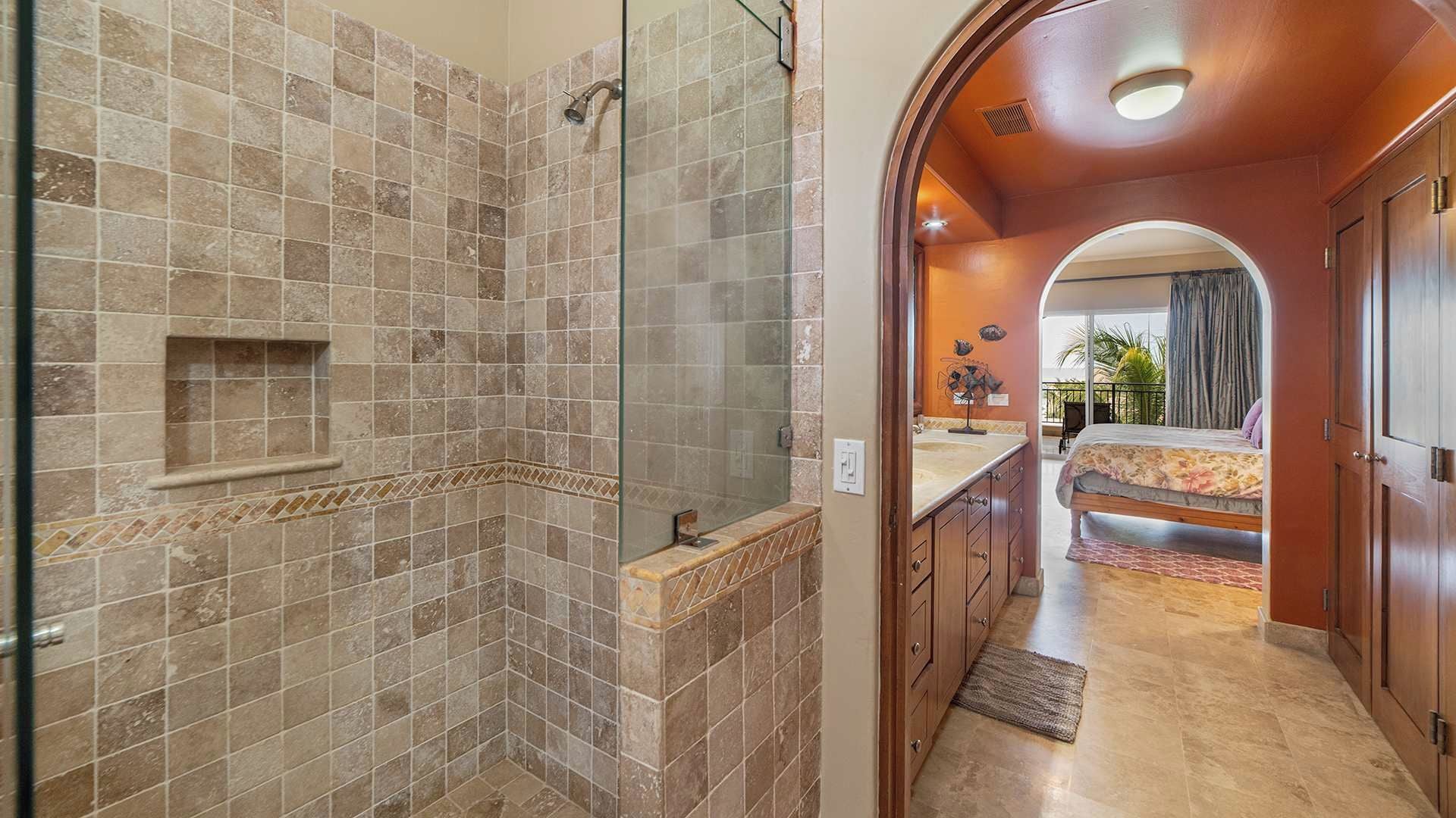 A bathroom with a shower, a vanity, and a bed in the adjacent room.