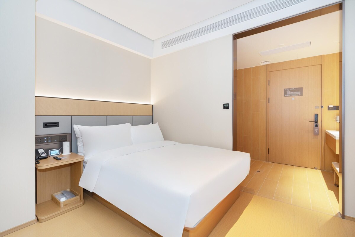 A modern hotel room with a neatly made bed, a bedside table with a phone and a lamp, and a wooden door leading to a bathroom.