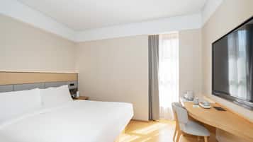 A hotel room with a large bed, a desk with a chair, a flat-screen TV, and a window with curtains.