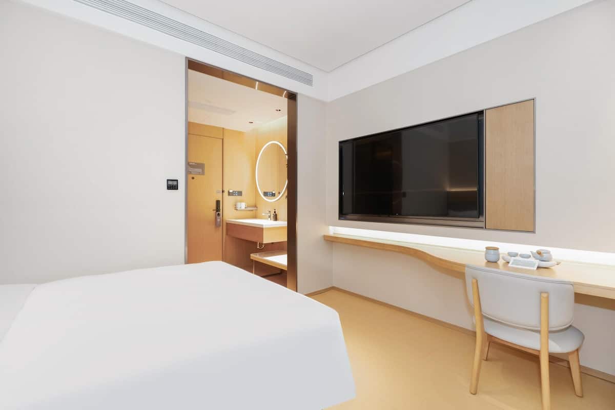 Ein modernes Hotelzimmer mit einem Bett, einem Schreibtisch mit Stuhl, einem großen Spiegel und einem an der Wand befestigten Fernseher.