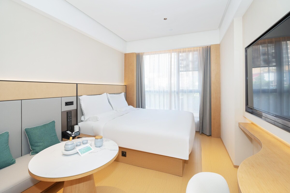 A modern hotel room with a large bed, a bedside table with a lamp, a small round table with a teapot and cups, and a window with curtains.