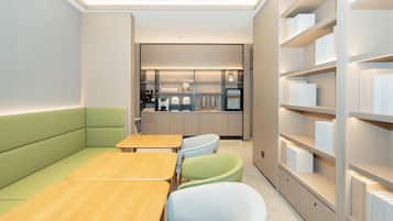 A modern, well-lit room with a green sofa, wooden tables, and light-colored chairs. A kitchen area with appliances and a refrigerator is visible in the background.