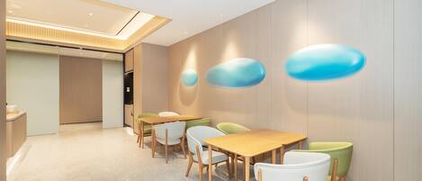 A modern dining area with wooden tables and chairs, a light-colored floor, and a wall with abstract blue shapes.