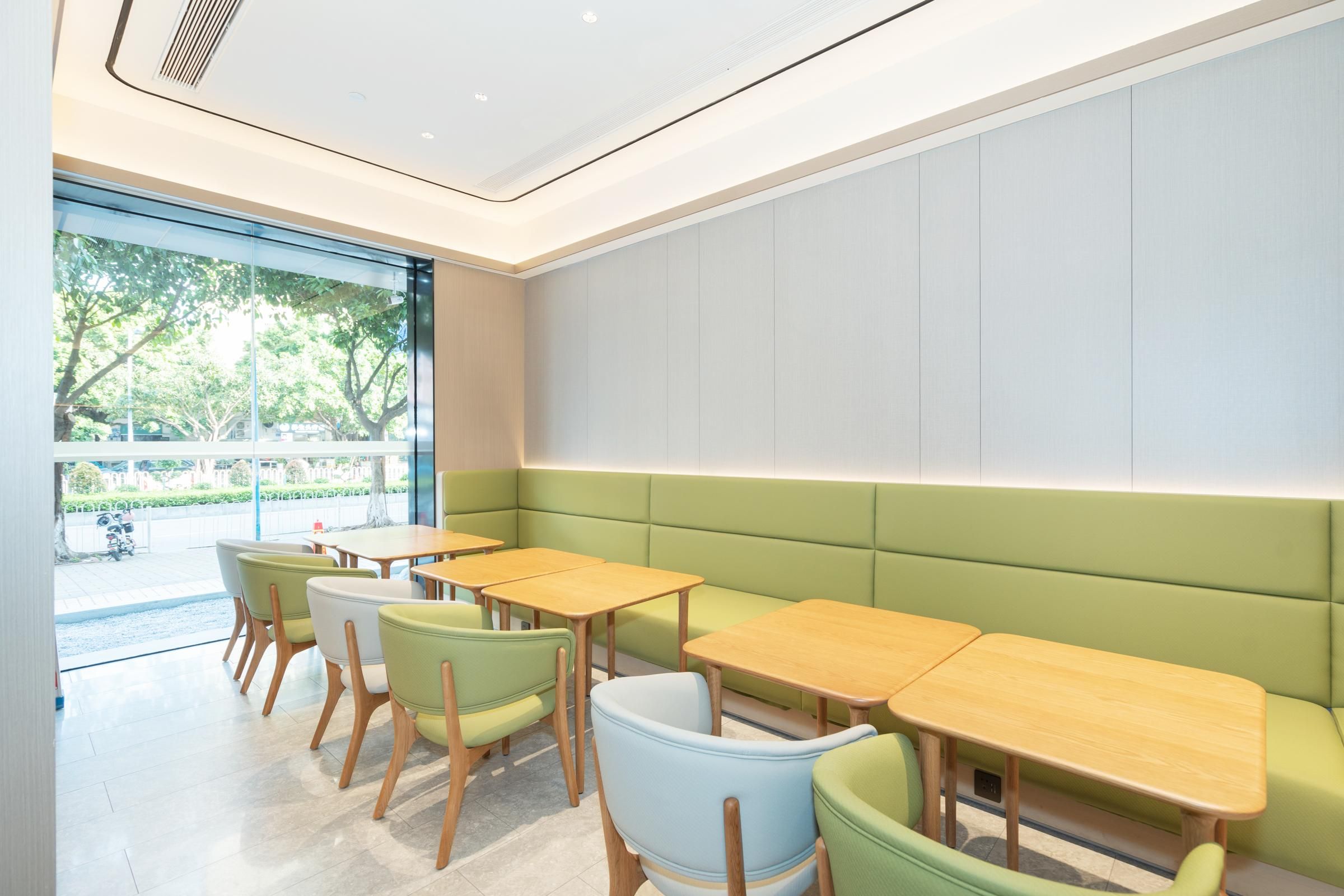 A modern cafe with green cushioned seating, wooden tables, and large windows offering a view of the outdoors.