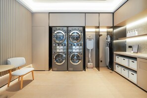 A modern laundry room with four stacked washing machines, a bench, a table, and a shelf with storage bins. - Ji Hotel Guangzhou Fangcun Huadiwan (Guangzhou)