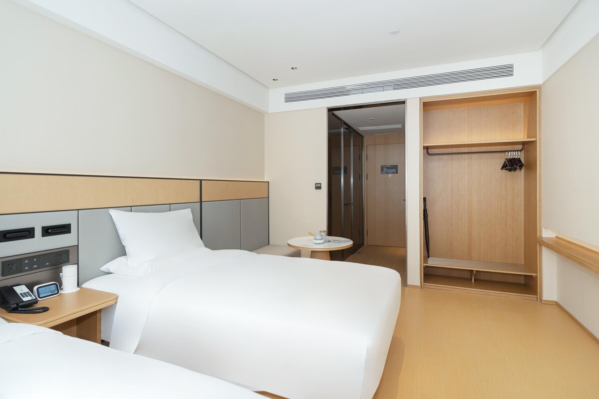 A hotel room with a large bed, bedside table with a phone and cup, a small round table with a cup, and a wooden wardrobe.