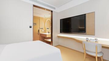 A modern hotel room with a bed, a desk with a chair, a large mirror, and a TV mounted on the wall.