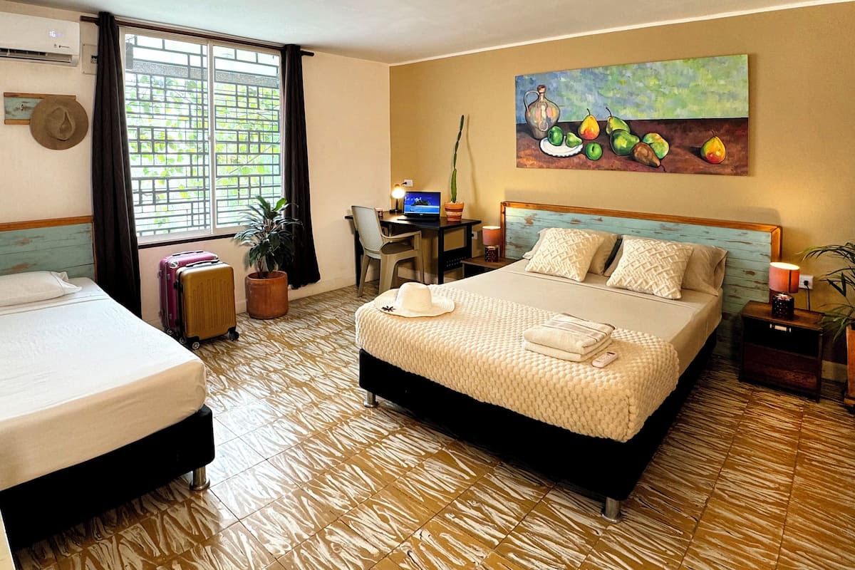A hotel room with two beds, a desk with a laptop, a painting on the wall, and a window with curtains.