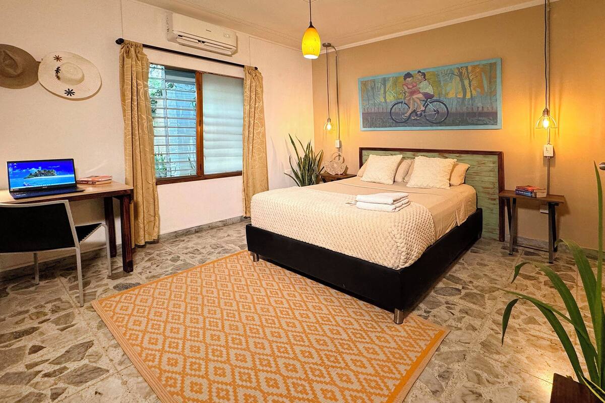 A bedroom with a bed, a desk with a laptop, a painting on the wall, and a potted plant.