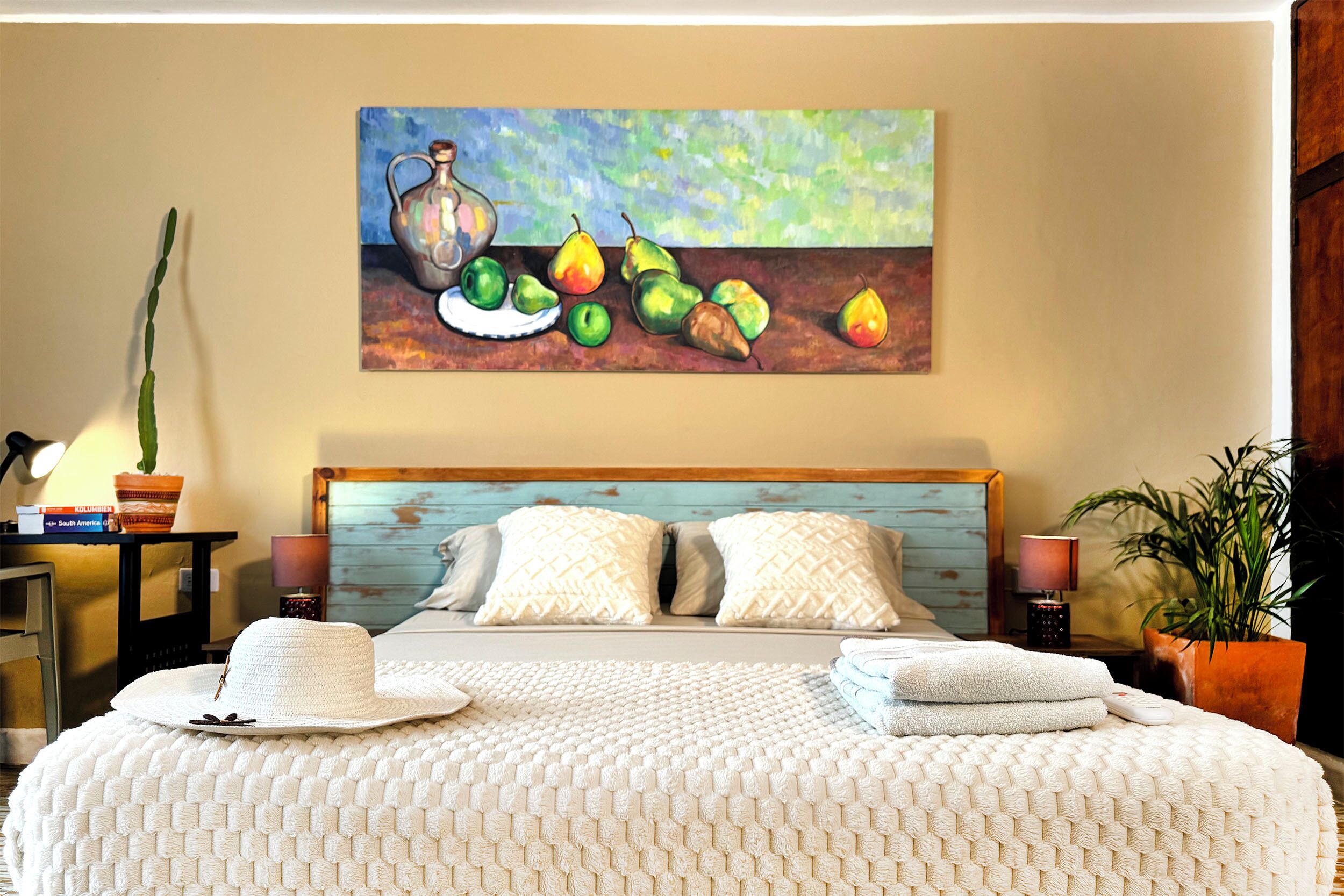 A bedroom with a bed, pillows, a painting, a lamp, and a plant.