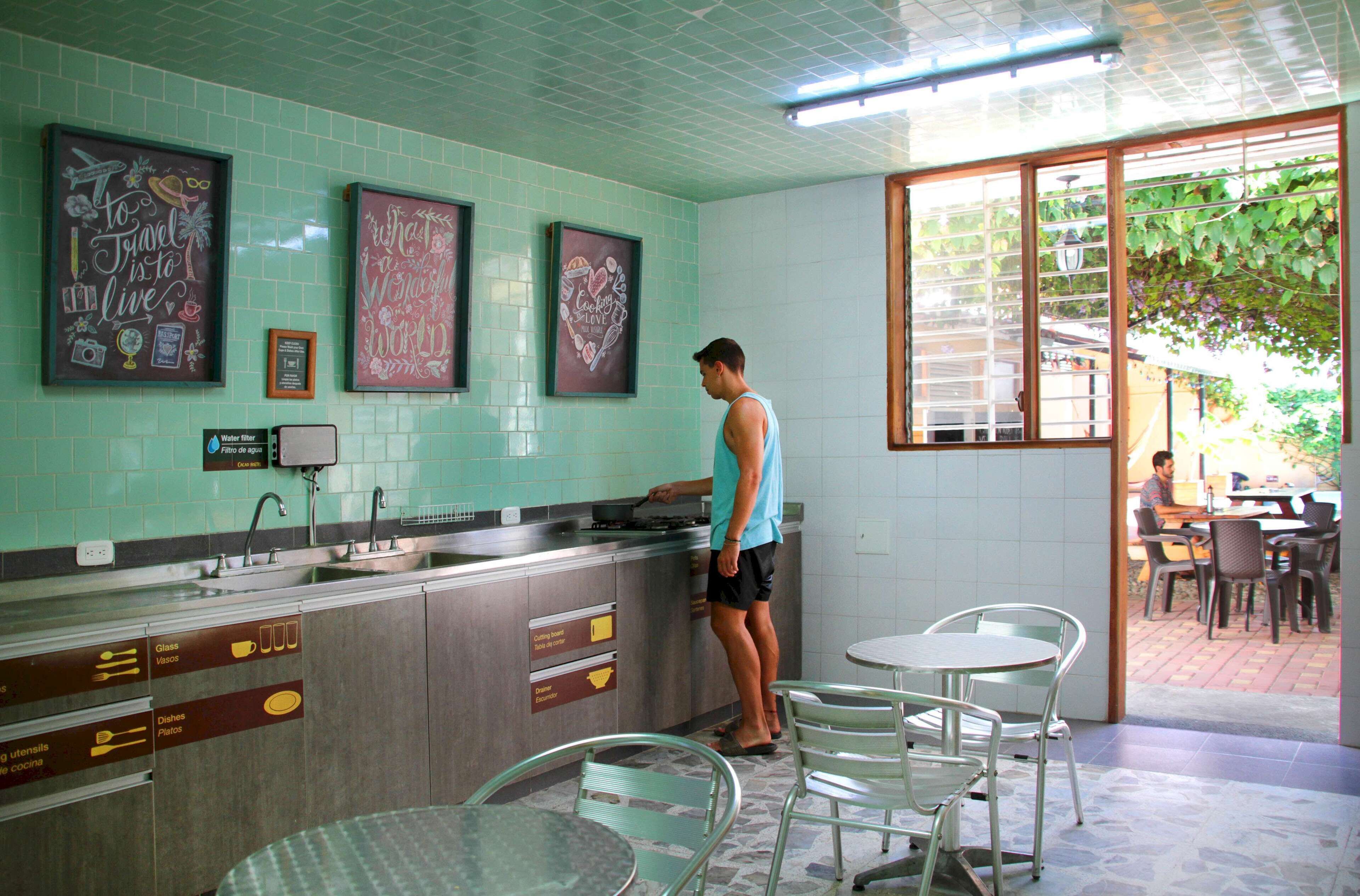 A kitchen with a person standing at the counter, a dining area with chairs and a table, and a view of the outdoors through a window.