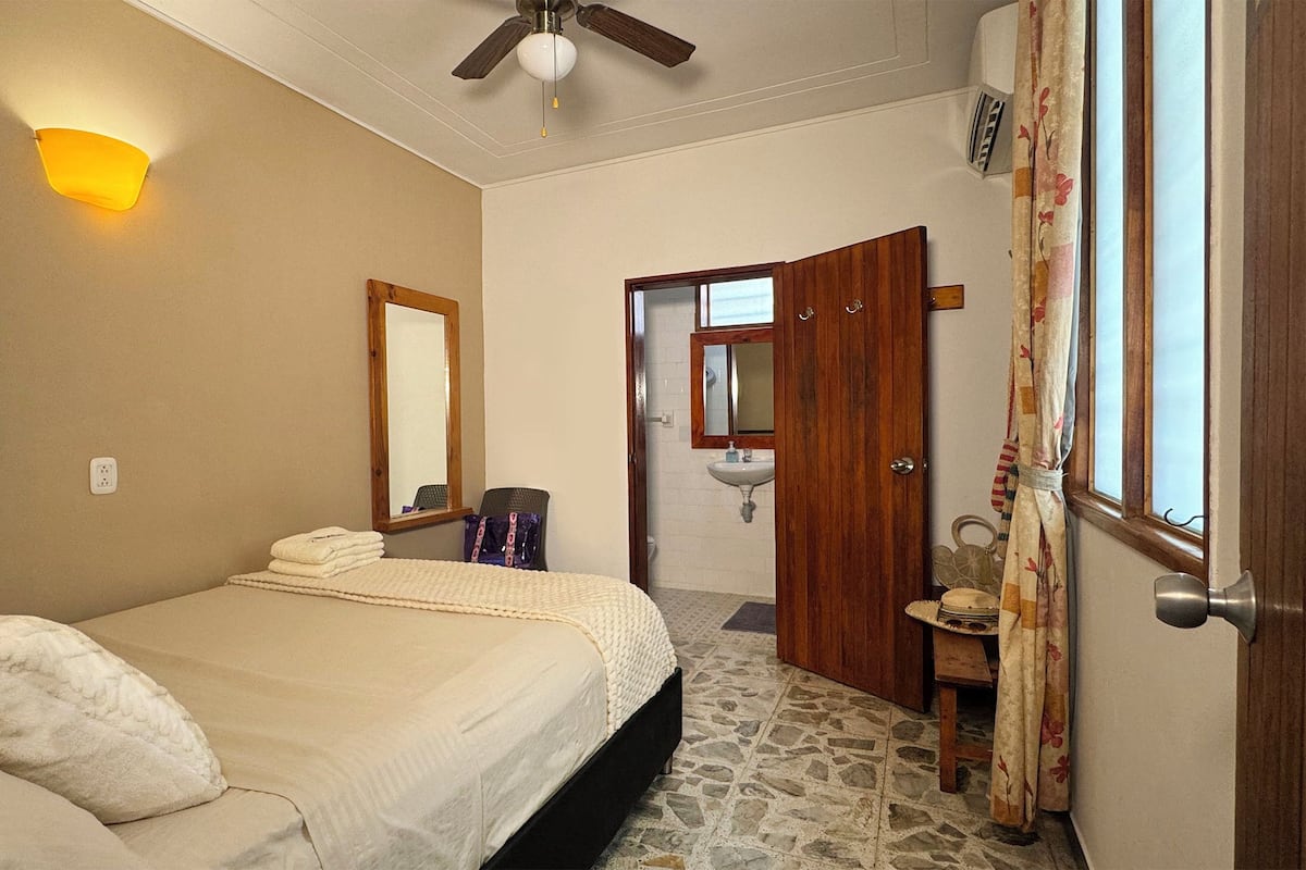 A bedroom with a bed, a ceiling fan, a mirror, a bathroom with a sink, and a window with curtains.