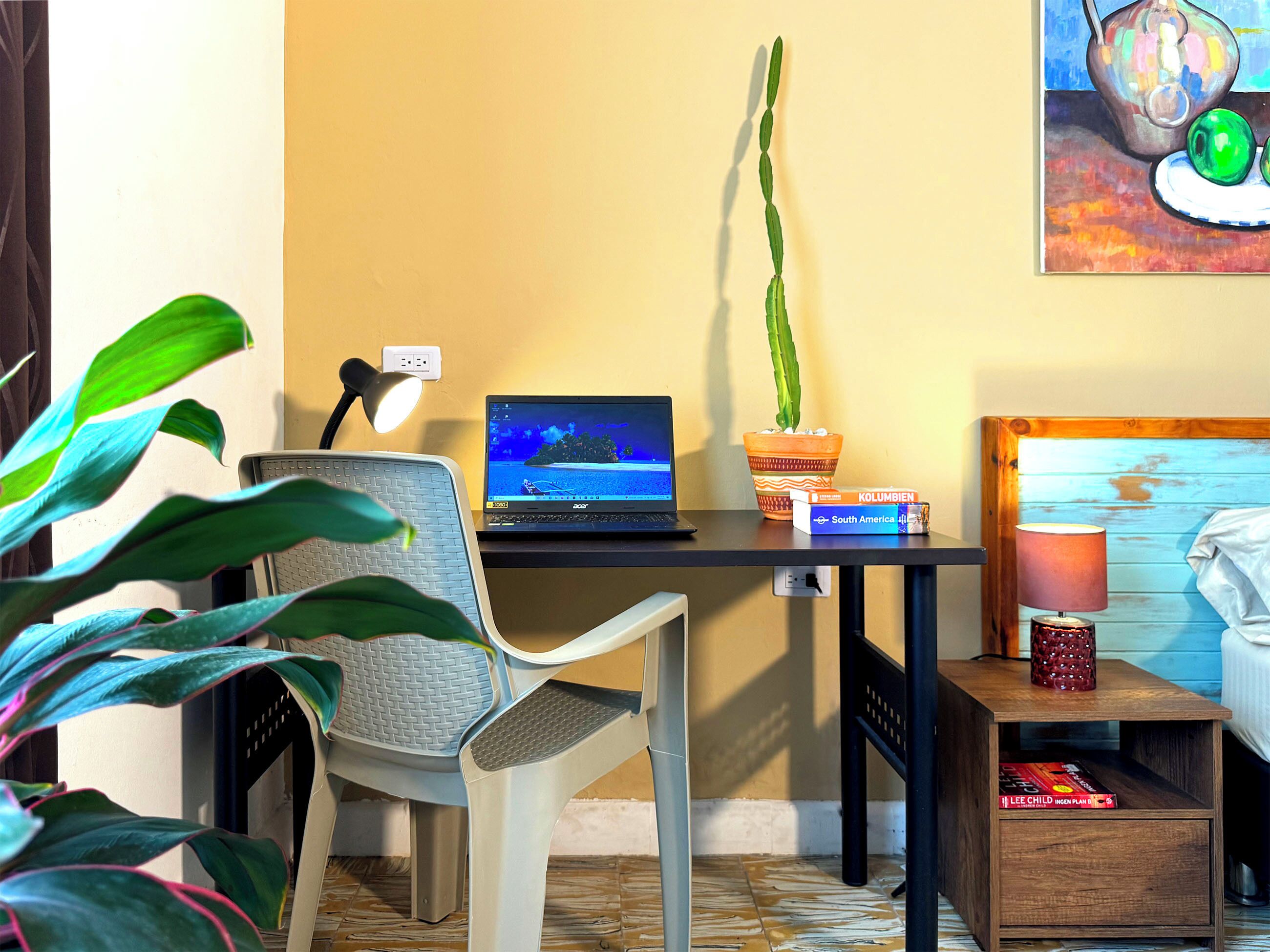 Una habitación con una laptop sobre un escritorio, una planta en maceta, una silla, una cama con lámpara y una estantería.