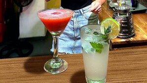 Ein Barkeeper bereitet ein Getränk an einer Bar zu, im Hintergrund eine Kreidetafel mit der Speisekarte und verschiedene Flaggen.