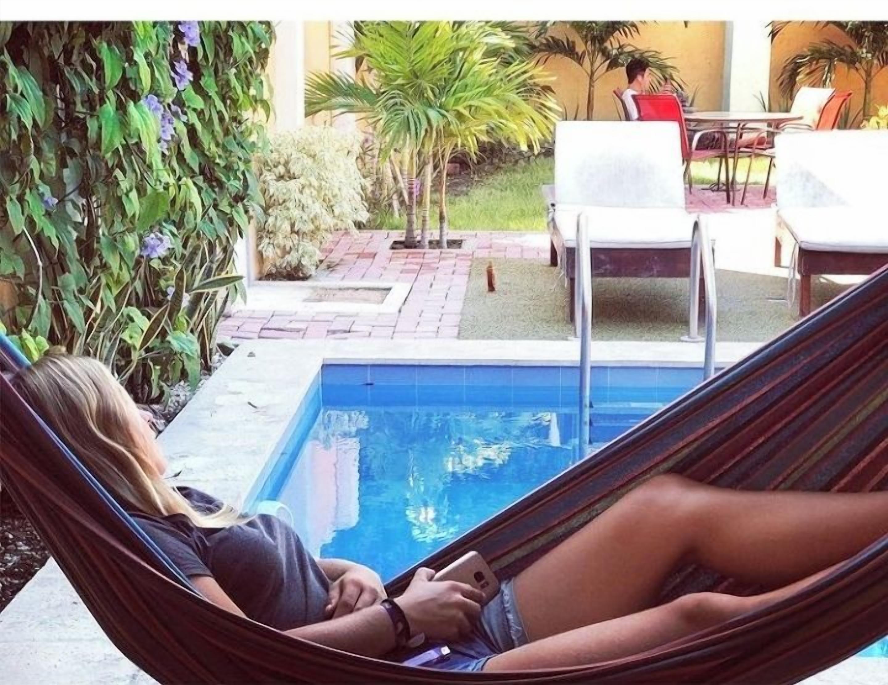 A person relaxing in a hammock by a pool with a garden and outdoor seating area in the background.
