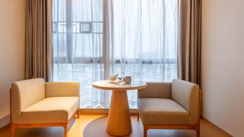 A modern room with two sofas facing a round wooden table, a large window with sheer curtains, and a view of a cityscape.