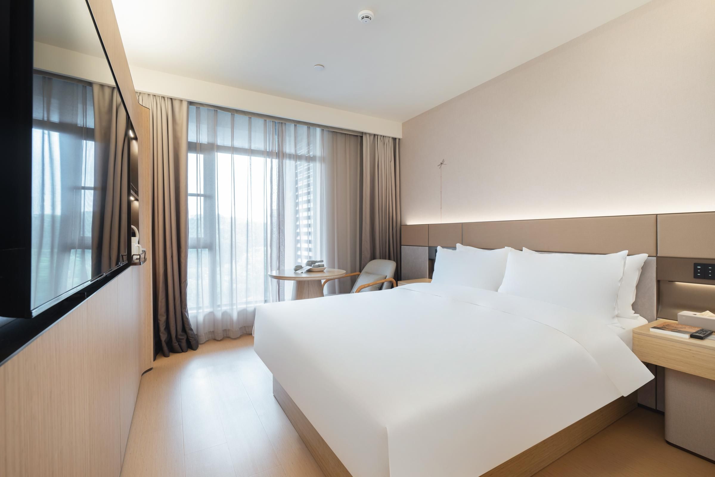 A modern hotel room with a large bed, a bedside table with a lamp, a small table with a chair, and a TV mounted on the wall.