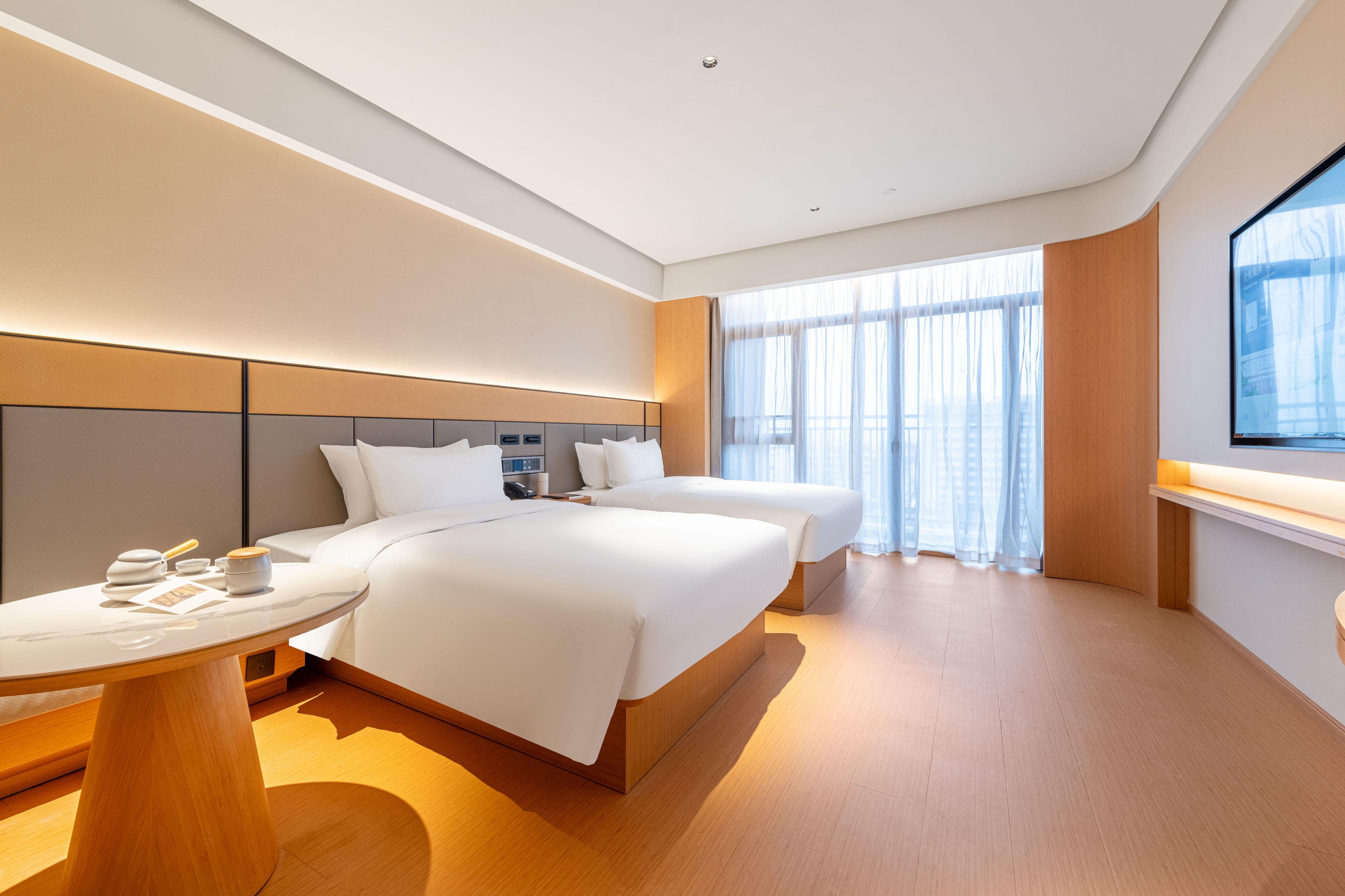 A modern hotel room with two beds, a round table, and a large window with sheer curtains.