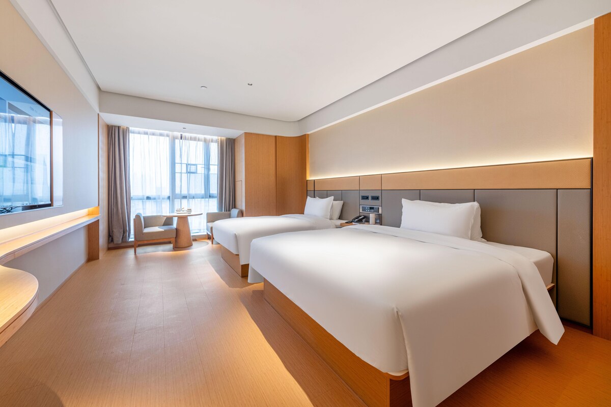 A modern hotel room with two beds, a desk, and a view of the city.