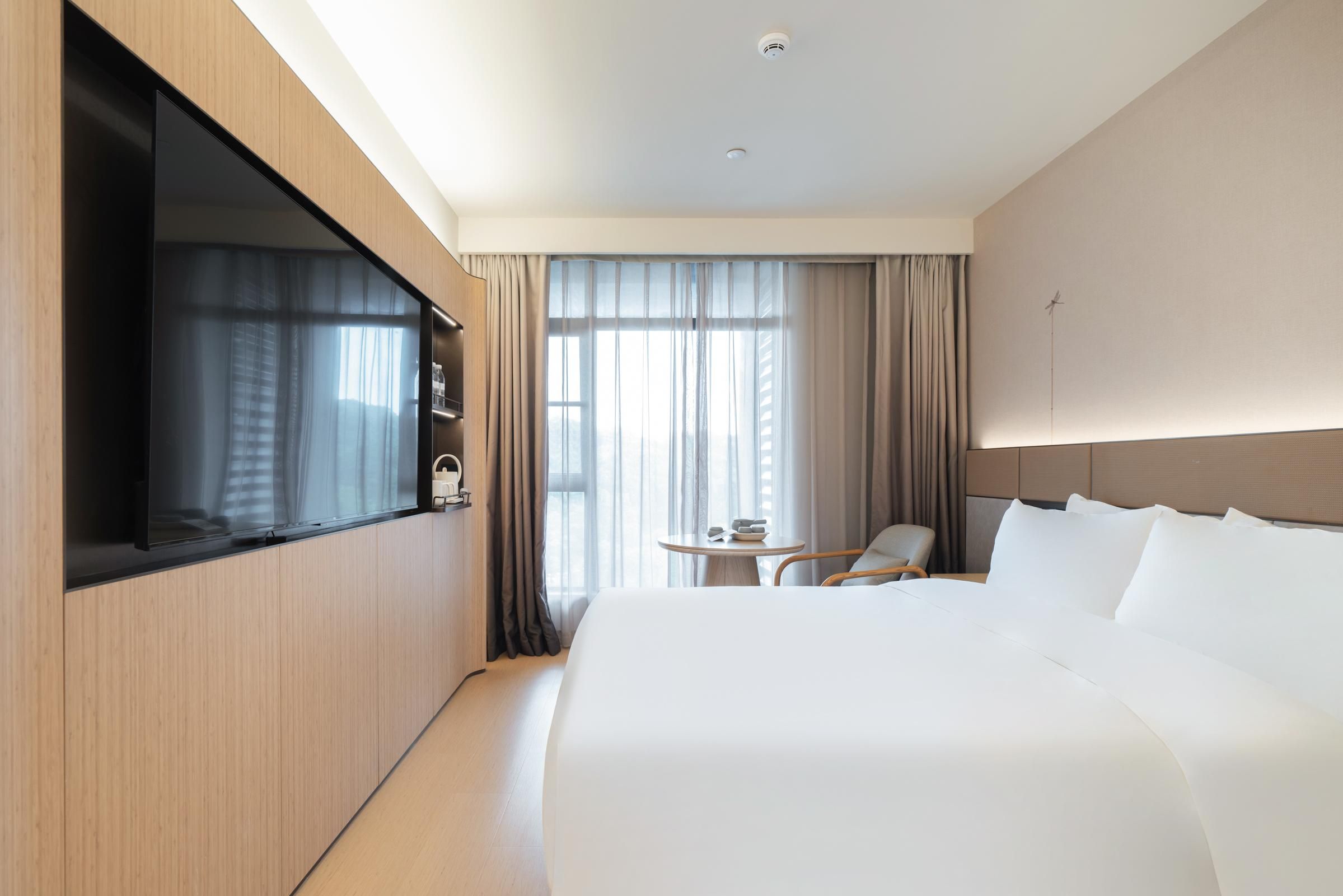 A hotel room with a large bed, a TV mounted on the wall, a small table with a chair, and a window with curtains.