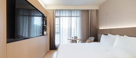 A hotel room with a large bed, a TV mounted on the wall, a small table with a chair, and a window with curtains.