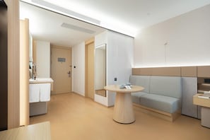 A modern hotel room with a round wooden table, a light blue sofa, a small round table with a mirror, and a bathroom with a sink and towel rack. - Ji Hotel Guangzhou Xintang South Station (Guangzhou)