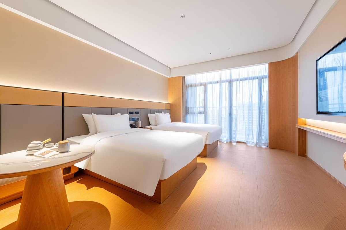 A modern hotel room with two beds, a round table, and a large window with sheer curtains.