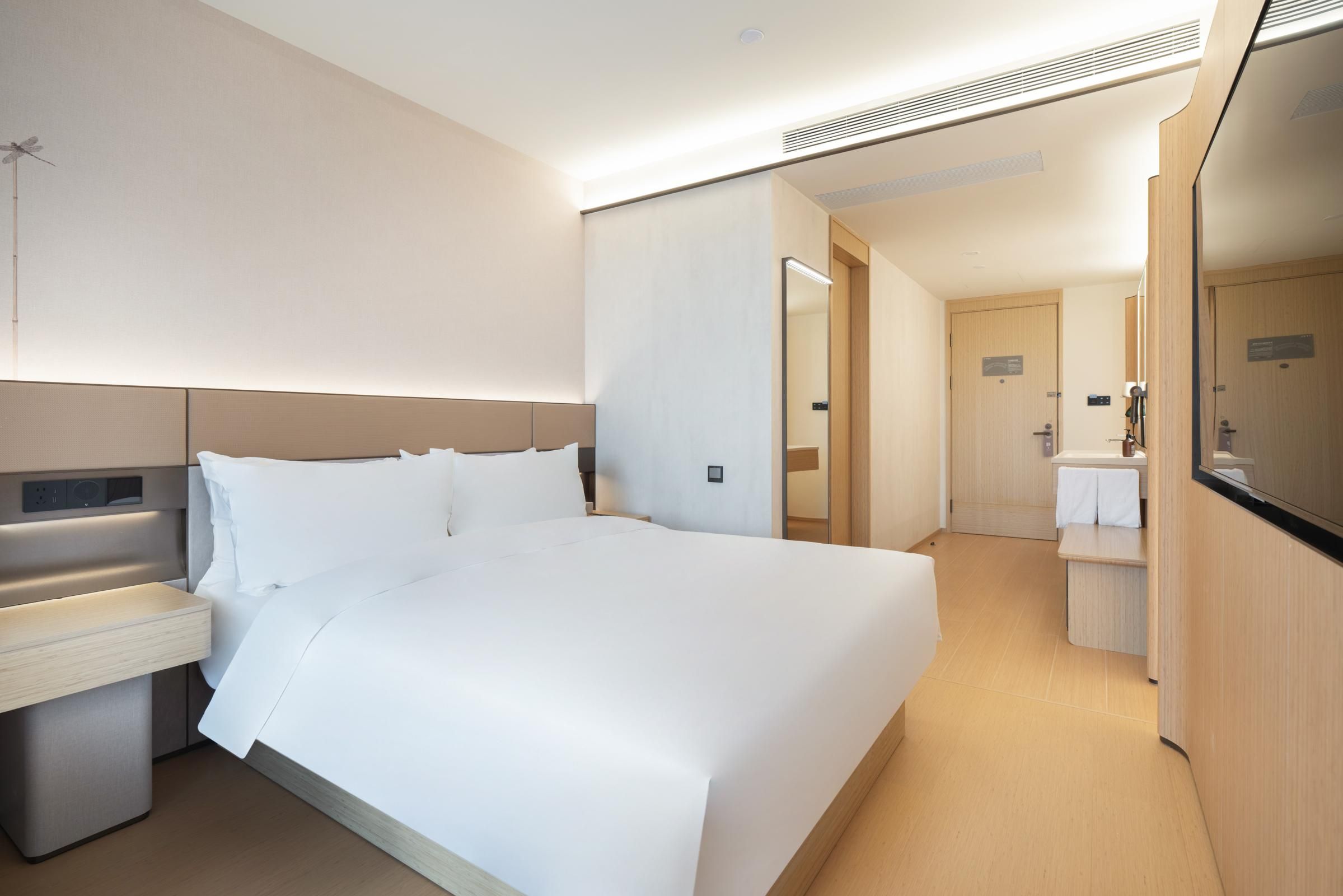 Ein modernes Hotelzimmer mit einem großen Bett, Nachttischen, einem Spiegel und einem kleinen Badezimmer mit Waschbecken und Handtuchhalter.
