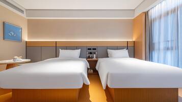 A hotel room with two beds, a bedside table with a lamp, a small table with a cup and saucer, and a painting on the wall.