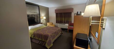 A hotel room with a bed, a chair, a desk with a lamp, a wall-mounted air conditioner, and a painting.