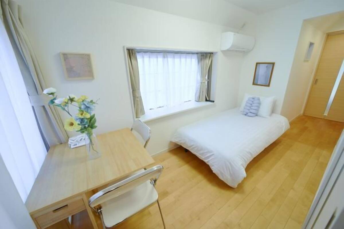 A small, clean room with a bed, a desk, a chair, a window with curtains, and a wall-mounted air conditioner.