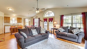 A spacious living area with two grey sofas, a coffee table, and a dining table with chairs. There is a kitchen in the background with wooden cabinets and a window with curtains. - 3 Mi to Arkansas River: Rural Family Home w/ Yard (Spiro)