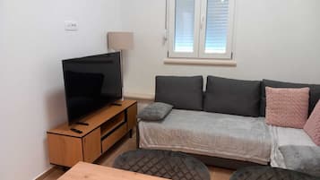 En lille, moderne stue med et fladskærms-tv på et træstel, en grå sofa med puder, et lille bord og et vindue med persienner.