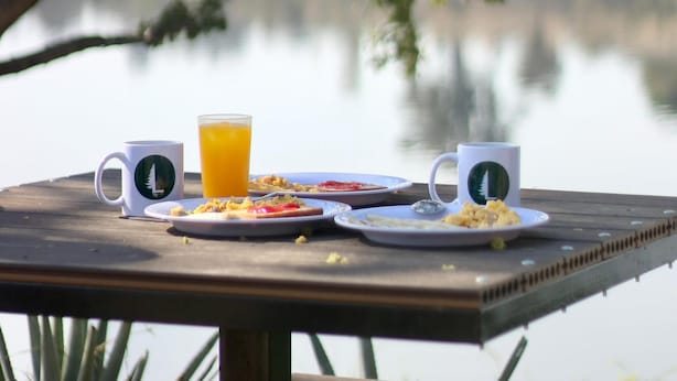 Una mesa de madera con alimentos para el desayuno y vasos de Starbucks, con vistas a un lago tranquilo.