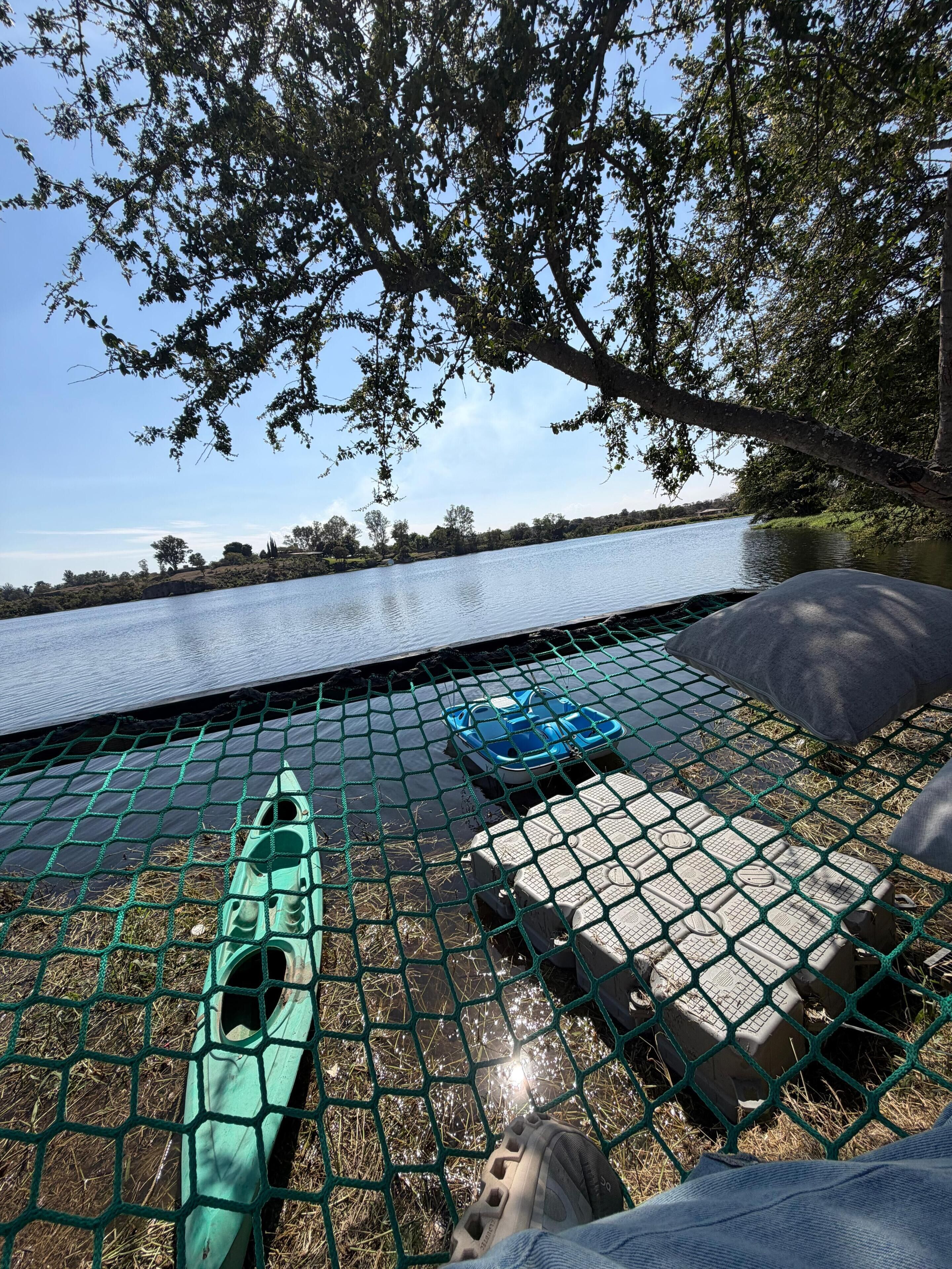 Una hamaca colocada en un espacio verde con vista a un lago tranquilo y árboles al fondo.
