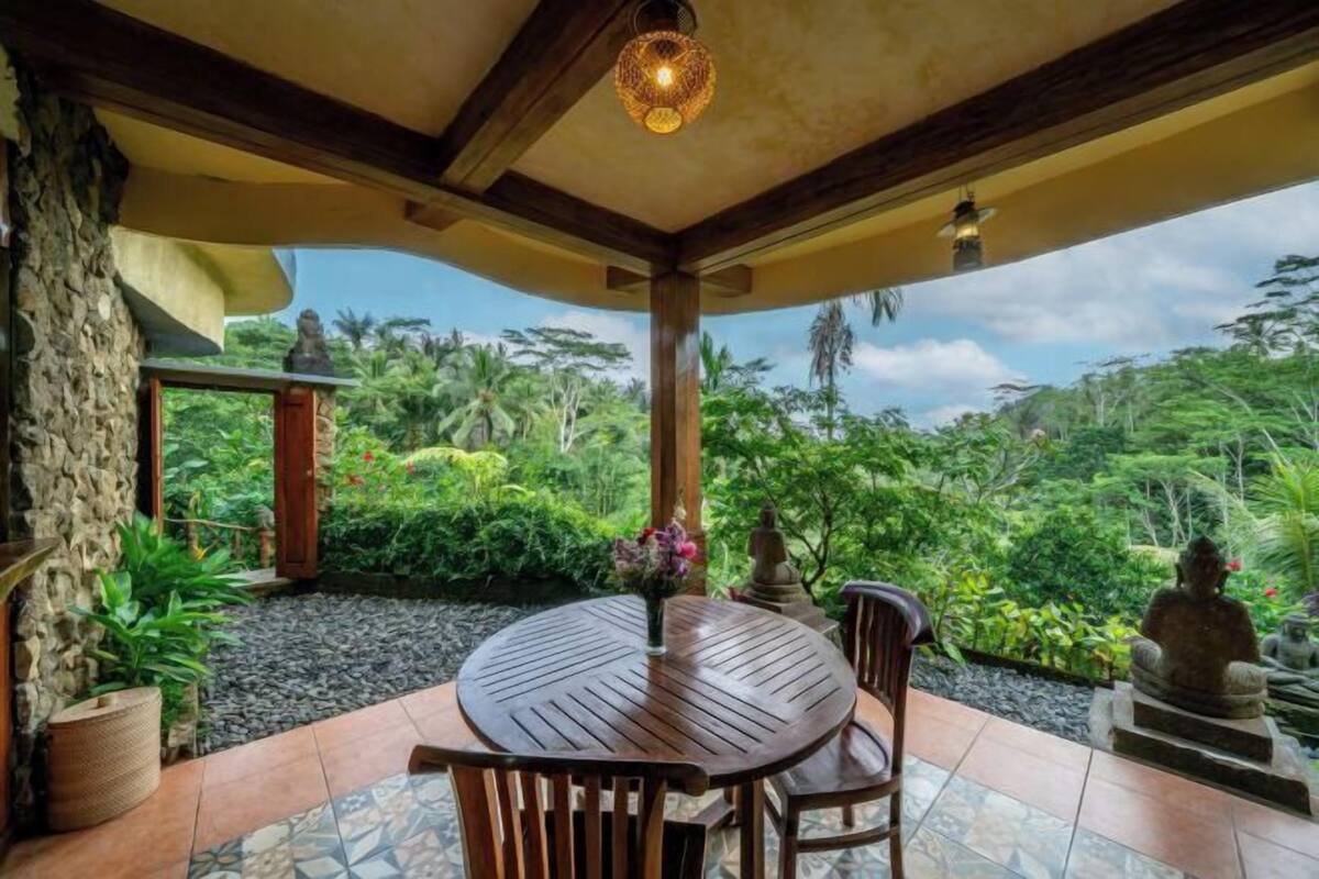 A round wooden table with chairs, a stone wall, and a view of lush greenery.