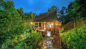 A rustic wooden house with a thatched roof, surrounded by lush greenery and a stone pathway leading to the entrance. - Yoga Maya Villa (Bali)