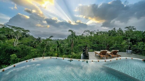 A swimming pool with lounge chairs, surrounded by lush greenery and a cloudy sky. - Yoga Maya Villa (Bali)