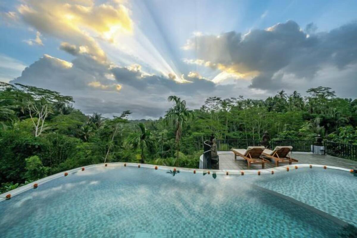 A swimming pool with lounge chairs, surrounded by lush greenery and a cloudy sky.