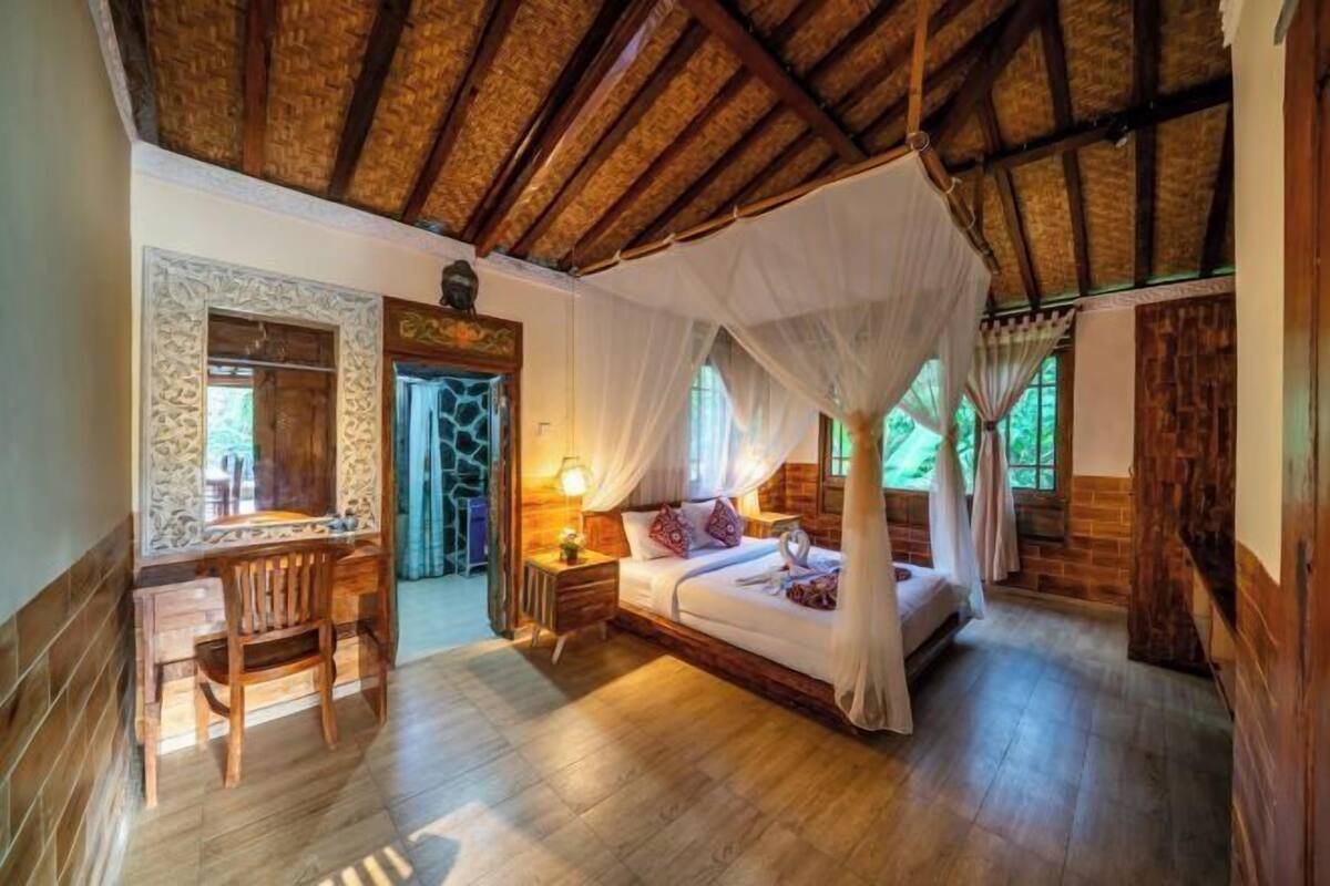 A room with a bed, a desk, and a chair, with a wooden ceiling and a stone wall.