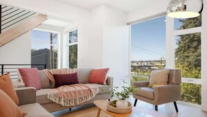 En moderne stue med sofa, lænestol og sofabord, store vinduer med udsigt samt en trappe.