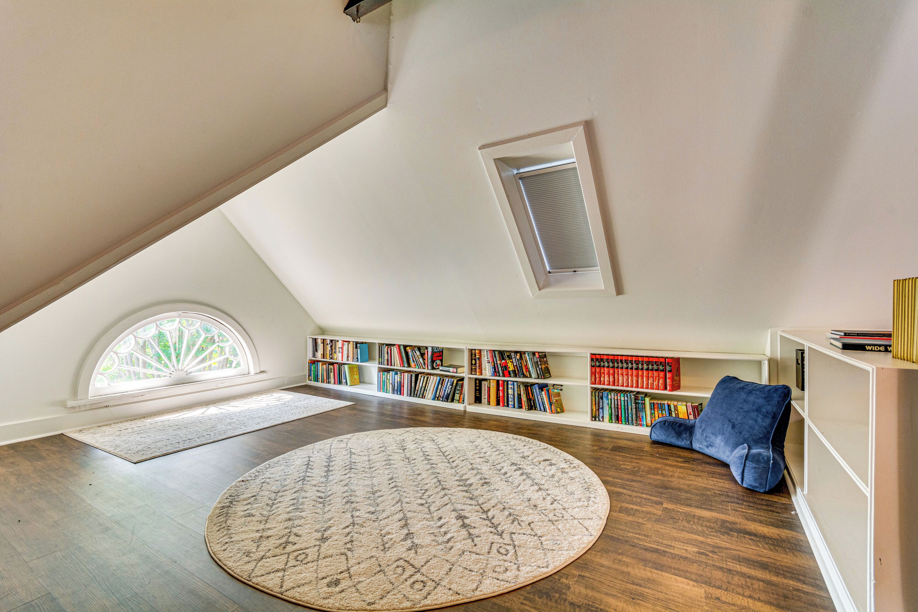 Un amplio cuarto ático con techo inclinado, un tragaluz, un tapete redondo grande, un sillón azul y una estantería llena de libros.
