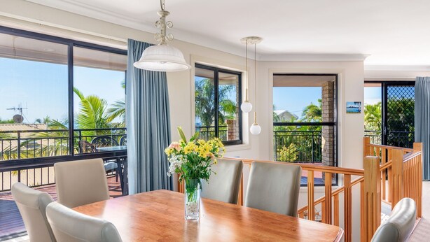 A dining area with a wooden table and chairs, a vase of flowers, and large windows offering a view of the outdoors.